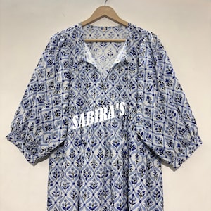 May include: A white and blue patterned dress with short sleeves and a V-neck. The dress features a floral design and the text "SABIRA'S" in white. The dress is hanging on a wooden hanger.