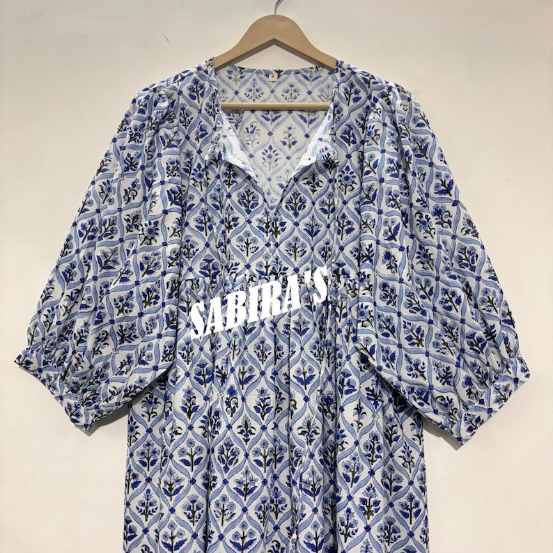 May include: A white and blue patterned dress with short sleeves and a V-neck. The dress features a floral design and the text "SABIRA'S" in white. The dress is hanging on a wooden hanger.