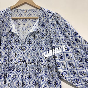 May include: A long-sleeved, white and blue floral patterned dress. The dress features a V-neckline and a gathered waist. The fabric has a repeating floral design in shades of blue and black. The dress is hanging on a wooden hanger.