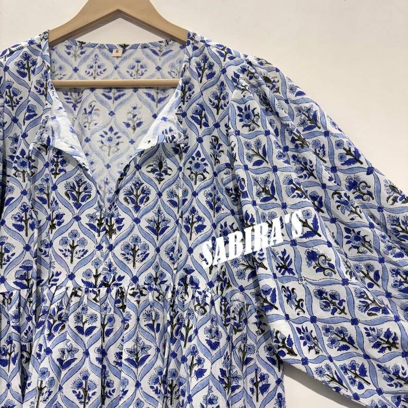 May include: A long-sleeved, white and blue floral patterned dress. The dress features a V-neckline and a gathered waist. The fabric has a repeating floral design in shades of blue and black. The dress is hanging on a wooden hanger.