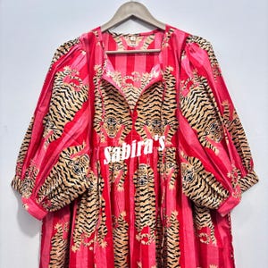 May include: A long-sleeved dress featuring a vibrant red and pink vertical striped pattern with a repeating tiger print throughout. The garment has a round neckline, gathered puff sleeves with cuffs, and the text Sabira's printed in white.