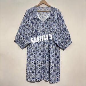 May include: A blue and white patterned dress with short sleeves and a V-neck. The dress features a floral design and the text "SABIRA'S" across the front. The dress is hanging on a wooden hanger.