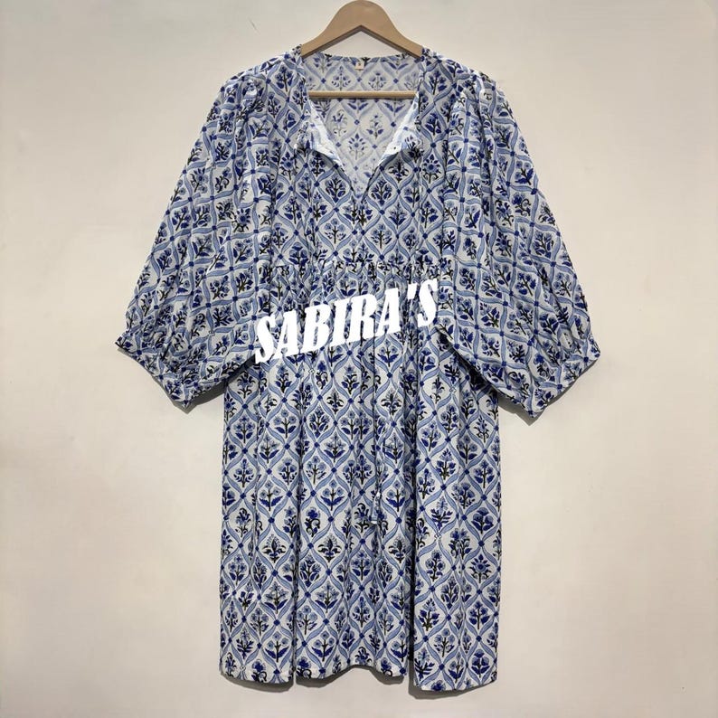 May include: A blue and white patterned dress with short sleeves and a V-neck. The dress features a floral design and the text "SABIRA'S" across the front. The dress is hanging on a wooden hanger.