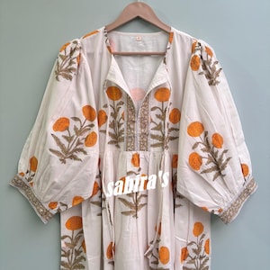 Puede incluir: Un vestido blanco con estampados florales naranjas y detalles marrones. El vestido presenta un escote en V, mangas largas y un corte holgado. El texto "sabita's" es visible en la parte delantera. Cuelga de una percha de madera.