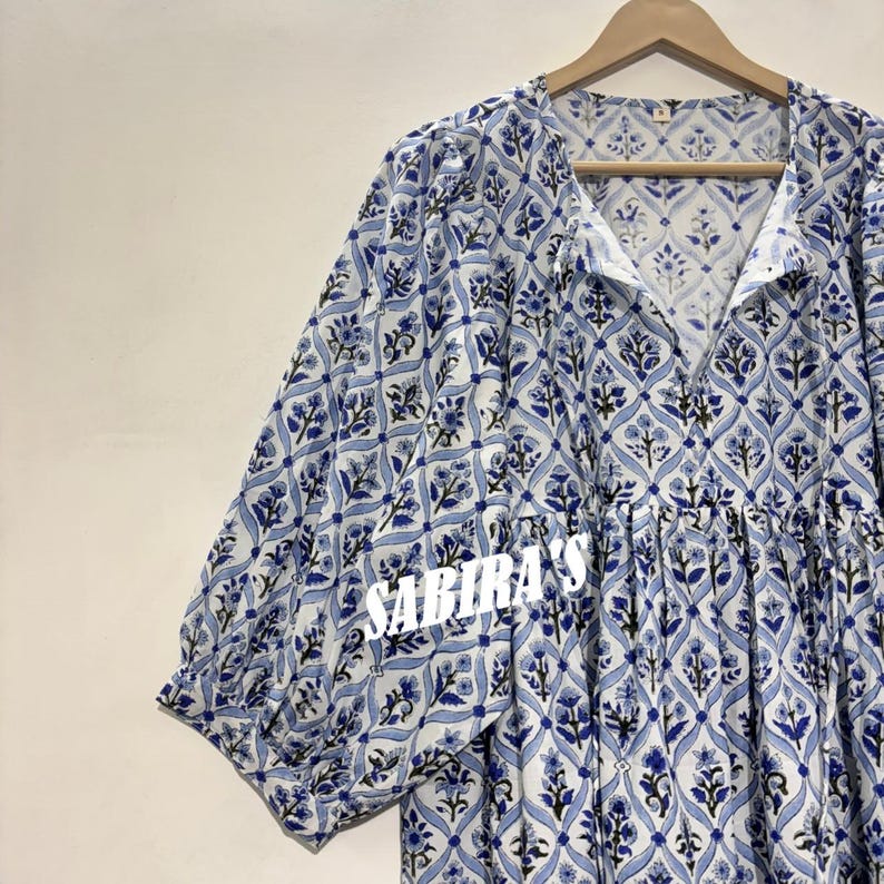 May include: A light blue and white patterned dress with short sleeves. The dress features a floral design and the text "SABIRA'S" in white. The dress is hanging on a wooden hanger.