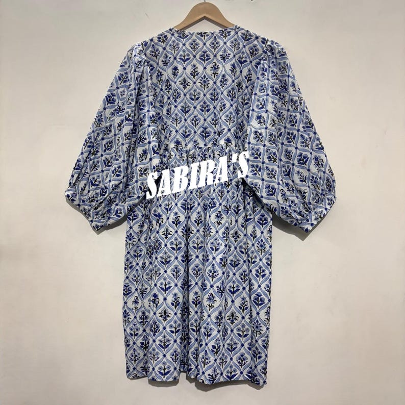 May include: A blue and white patterned dress with long, puffy sleeves. The dress features a floral design and the text "SABIRA'S" across the midsection. The dress is hanging on a wooden hanger.