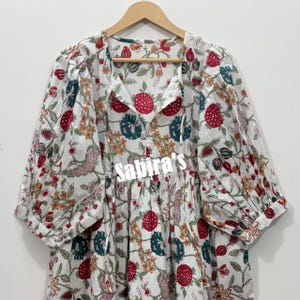 May include: A white blouse featuring a floral block print pattern in shades of red, green, and orange. The garment has long, voluminous sleeves with gathered cuffs, a V-neckline with tie closures, and the text Sabira's printed across the center.
