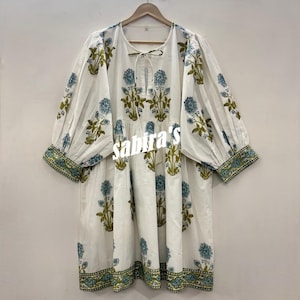Pu&ograve; includere: Abito tunica bianco con motivo floreale blu e verde oliva. L'abito ha maniche lunghe a sbuffo e un fiocco allo scollo. L'orlo e i polsini sono rifiniti con un bordo a fantasia. Il testo "sabira's" &egrave; visibile sul davanti.
