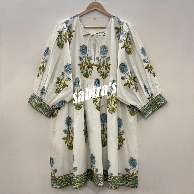 Pu&ograve; includere: Abito tunica bianco con motivo floreale blu e verde oliva. L'abito ha maniche lunghe a sbuffo e un fiocco allo scollo. L'orlo e i polsini sono rifiniti con un bordo a fantasia. Il testo "sabira's" &egrave; visibile sul davanti.