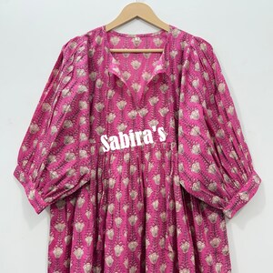 May include: A bright pink dress featuring a repeating floral pattern in light beige and dark outlines. The garment has a split V-neckline, voluminous three-quarter length sleeves with gathered cuffs, and the text Sabira's printed in white.