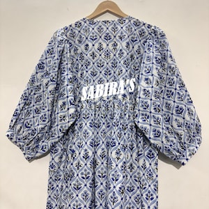 May include: A long-sleeved dress with a blue and white floral pattern. The dress has a gathered waist and the text "SABIRA'S" in white across the back. The sleeves are wide and gathered at the cuffs.