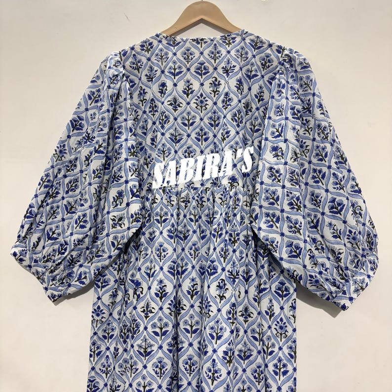 May include: A long-sleeved dress with a blue and white floral pattern. The dress has a gathered waist and the text "SABIRA'S" in white across the back. The sleeves are wide and gathered at the cuffs.