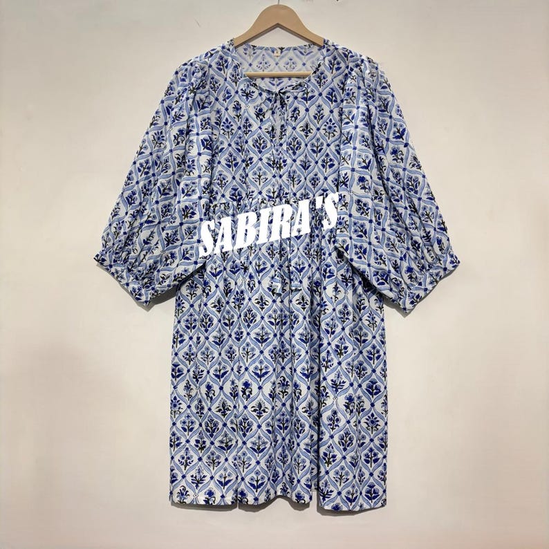 May include: A blue and white patterned dress with short sleeves and a round neckline. The dress features a floral design and the text "SABIRA'S" across the front. The dress is hanging on a wooden hanger.