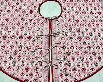 May include: A round, white and pink patterned tree skirt with a red border. The skirt has a center opening and a split front with ties. The floral pattern is repeated across the surface.