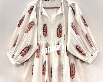 Puede incluir: Vestido blanco de manga larga con cuello en V y estampado floral en rojo, morado y verde. El vestido tiene una silueta suelta y está colgado en una percha de madera.
