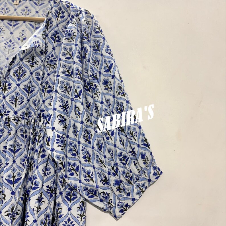 May include: A white and blue patterned garment with short sleeves. The fabric features a repeating floral design in shades of blue. The text "SABIRA'S" is visible in white on the right side of the image. The garment is hanging on a wooden hanger.