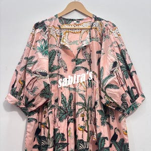 May include: A light pink tunic featuring a jungle-themed print with leopards, flamingos, toucans, and palm trees. The garment has three-quarter length puff sleeves, a split neckline with thin ties, and the word sabira's printed in white across the chest.
