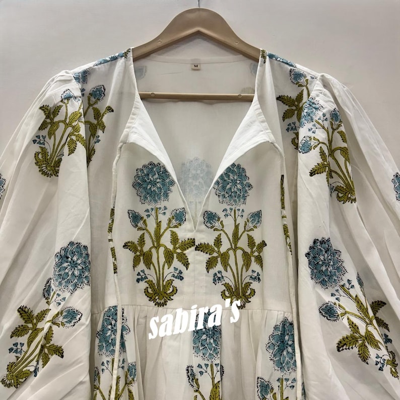 Pu&ograve; includere: Abito in cotone bianco con scollo a V e maniche lunghe a sbuffo. L'abito presenta un motivo floreale nei toni del blu e del verde. Il testo "sabira's" &egrave; visibile sul davanti.