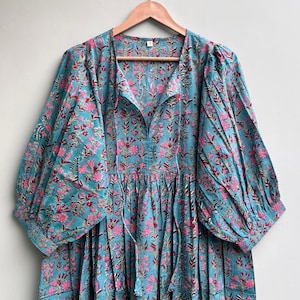 Puede incluir: Un vestido largo y fluido con un fondo turquesa y un estampado floral rosa. El vestido tiene mangas largas y abullonadas y un escote en V con cordón. El vestido está colgado de una percha de madera.