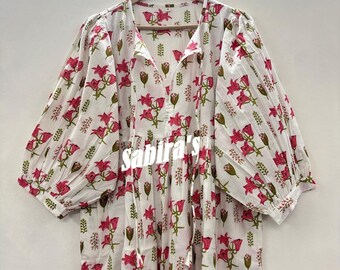 May include: White dress with a pink floral pattern and green accents. The dress has a V-neck, long sleeves, and a drawstring at the neckline. The dress is hanging on a wooden hanger.