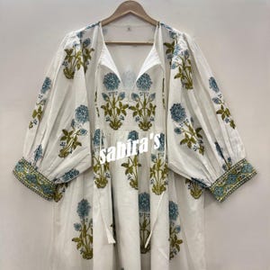 以下が含まれることがあります： Vネックと長袖の白いドレス。ドレスは青と緑の花柄で、中央に「sabira's」の文字が入っています。袖口は模様入りです。
