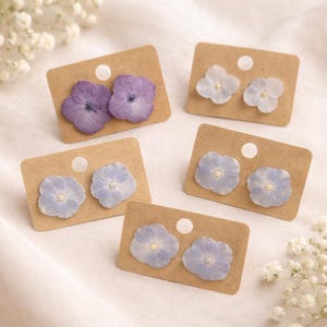 May include: Five pairs of floral earrings displayed on small, rectangular brown cards. The earrings feature pressed flowers in shades of purple and light blue. The cards have a small hole at the top for display.