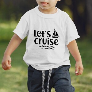 Peut inclure: T-shirt blanc avec l'inscription "Let's Cruise" en noir, orné d'un dessin de voilier au-dessus de vagues. Le t-shirt est porté par un enfant, suggérant un thème nautique ou de vacances.