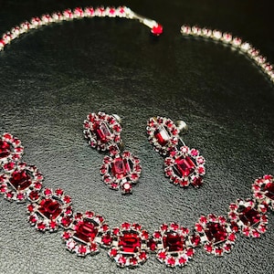 May include: A silver-toned necklace and matching pair of screw-back earrings featuring deep red, square-cut faceted stones surrounded by a halo of smaller, round, brilliant-cut red crystals set in a metallic frame with a polished finish.