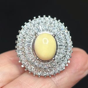 May include: A close-up of a ring featuring a large, oval, pale yellow gemstone at the center. The gemstone is surrounded by multiple concentric circles of small, clear, rectangular-cut stones, creating a sparkling halo effect. The ring is set on a light skin tone.