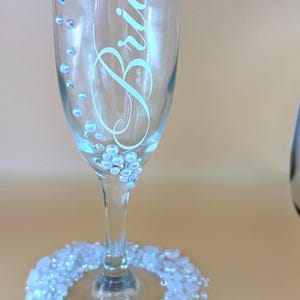 Puede incluir: Una copa de champ&aacute;n transparente con la palabra "Bride" en cursiva. El vaso est&aacute; adornado con pedrer&iacute;a iridiscente y una base de gemas blancas e iridiscentes. El dise&ntilde;o es elegante y adecuado para una boda o un evento nupcial.