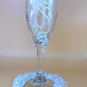 Puede incluir: Una copa de champ&aacute;n transparente con la palabra "Bride" en escritura cursiva. El vaso est&aacute; adornado con pedrer&iacute;a iridiscente y un racimo de perlas. La base est&aacute; rodeada por un anillo de cuentas blancas e iridiscentes.