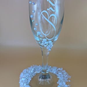 May include: A clear champagne flute with the word "Bride" in elegant script. The glass is adorned with small, sparkling embellishments and a base ringed with white beads, creating a celebratory and decorative design.