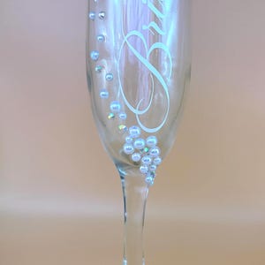 Puede incluir: Una copa de champ&aacute;n transparente con la palabra &laquo;Bride&raquo; en escritura blanca. El cristal est&aacute; decorado con perlas de imitaci&oacute;n y pedrer&iacute;a iridiscente. El tallo y la base son de cristal transparente.