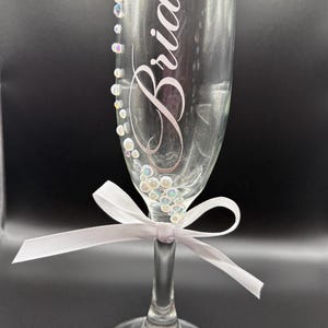 Puede incluir: Una flauta de champ&aacute;n transparente con la palabra "Bride" en cursiva. El vaso est&aacute; adornado con pedrer&iacute;a iridiscente y una cinta blanca atada alrededor del tallo. El vaso est&aacute; dise&ntilde;ado para una boda.
