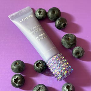 May include: A purple tube of Laneige Lip Glowy Balm with the text "Lip Glowy Balm Baume Brillant Pour Les Lèvres Blueberry Myrtille" on the label. The tube is surrounded by blueberries on a purple background.