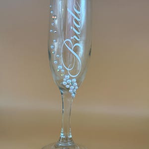 May include: A clear champagne flute with the word "Bride" in elegant script. The glass is adorned with small, pearl-like embellishments. The stem and base are also clear, creating a delicate and celebratory aesthetic.