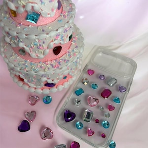 Puede incluir: Una funda de teléfono transparente decorada con varios diamantes de imitación de plástico de colores en formas de corazón, redondas y rectangulares, junto a un adorno de pastel de espuma escalonado con glaseado blanco, chispas y gemas.