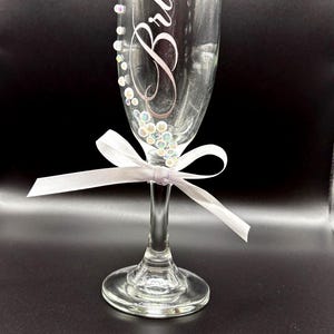 Puede incluir: Una flauta de champ&aacute;n transparente con la palabra "Bride" en escritura cursiva. El vaso est&aacute; adornado con cuentas iridiscentes y una cinta blanca atada en un lazo. El borde del vaso tambi&eacute;n est&aacute; decorado con peque&ntilde;as cuentas. Un art&iacute;culo tem&aacute;tico de boda.