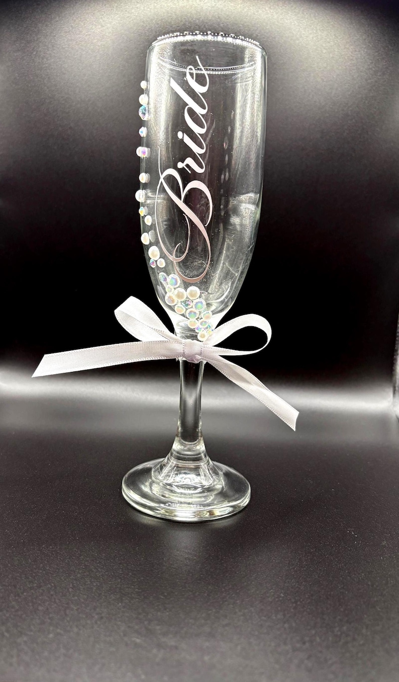 Puede incluir: Una flauta de champ&aacute;n transparente con la palabra "Bride" en escritura cursiva. El vaso est&aacute; adornado con cuentas iridiscentes y una cinta blanca atada en un lazo. El borde del vaso tambi&eacute;n est&aacute; decorado con peque&ntilde;as cuentas. Un art&iacute;culo tem&aacute;tico de boda.