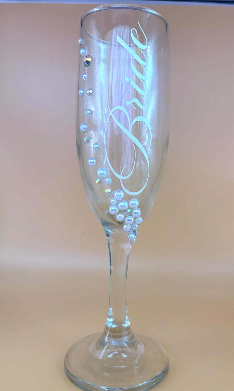 Puede incluir: Una copa de champ&aacute;n transparente con la palabra &laquo;Bride&raquo; en escritura blanca. El cristal est&aacute; decorado con perlas de imitaci&oacute;n y pedrer&iacute;a iridiscente. El tallo y la base son de cristal transparente.