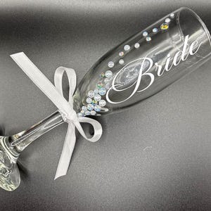 Puede incluir: Una copa de champ&aacute;n transparente con la palabra "Bride" en escritura blanca. El vaso est&aacute; adornado con pedrer&iacute;a iridiscente y una cinta blanca atada con un lazo. Un art&iacute;culo tem&aacute;tico de boda.