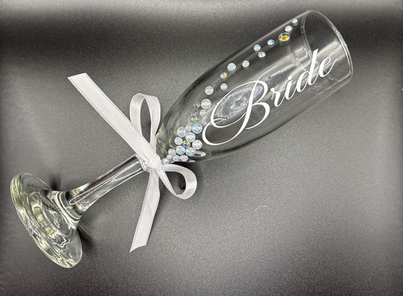 Puede incluir: Una copa de champ&aacute;n transparente con la palabra "Bride" en escritura blanca. El vaso est&aacute; adornado con pedrer&iacute;a iridiscente y una cinta blanca atada con un lazo. Un art&iacute;culo tem&aacute;tico de boda.