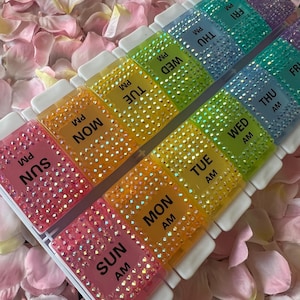 Puede incluir: Un organizador de pastillas para siete d&iacute;as con una caja de pl&aacute;stico transparente y compartimentos coloridos. Cada compartimento est&aacute; decorado con diamantes de imitaci&oacute;n y etiquetado con los d&iacute;as de la semana y AM o PM.
