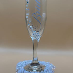 Puede incluir: Una copa de champ&aacute;n transparente con la palabra "Bride" en escritura azul claro. El vaso est&aacute; decorado con peque&ntilde;as cuentas iridiscentes y un grupo de adornos blancos y transparentes en la base. El vaso se asienta sobre un lecho de cuentas blancas y transparentes.