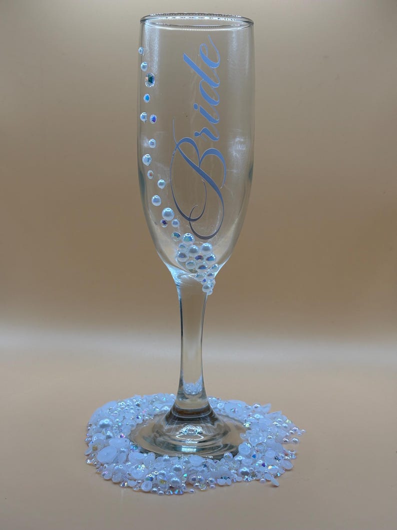 Puede incluir: Una copa de champ&aacute;n transparente con la palabra "Bride" en escritura azul claro. El vaso est&aacute; decorado con peque&ntilde;as cuentas iridiscentes y un grupo de adornos blancos y transparentes en la base. El vaso se asienta sobre un lecho de cuentas blancas y transparentes.