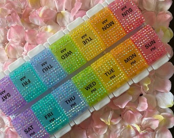 May include: A rainbow pill organiser with compartments for each day of the week, labelled with AM and PM. The organiser is decorated with rhinestones.