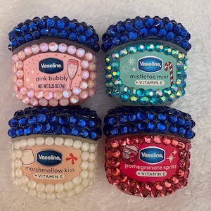 Peut inclure: Quatre pots de Vaseline pour les lèvres décorés. Chaque pot est orné de strass colorés et présente une saveur différente : pink bubbly, mistletoe mint, marshmallow kiss et pomegranate spritz. Les pots sont surmontés de strass bleus.
