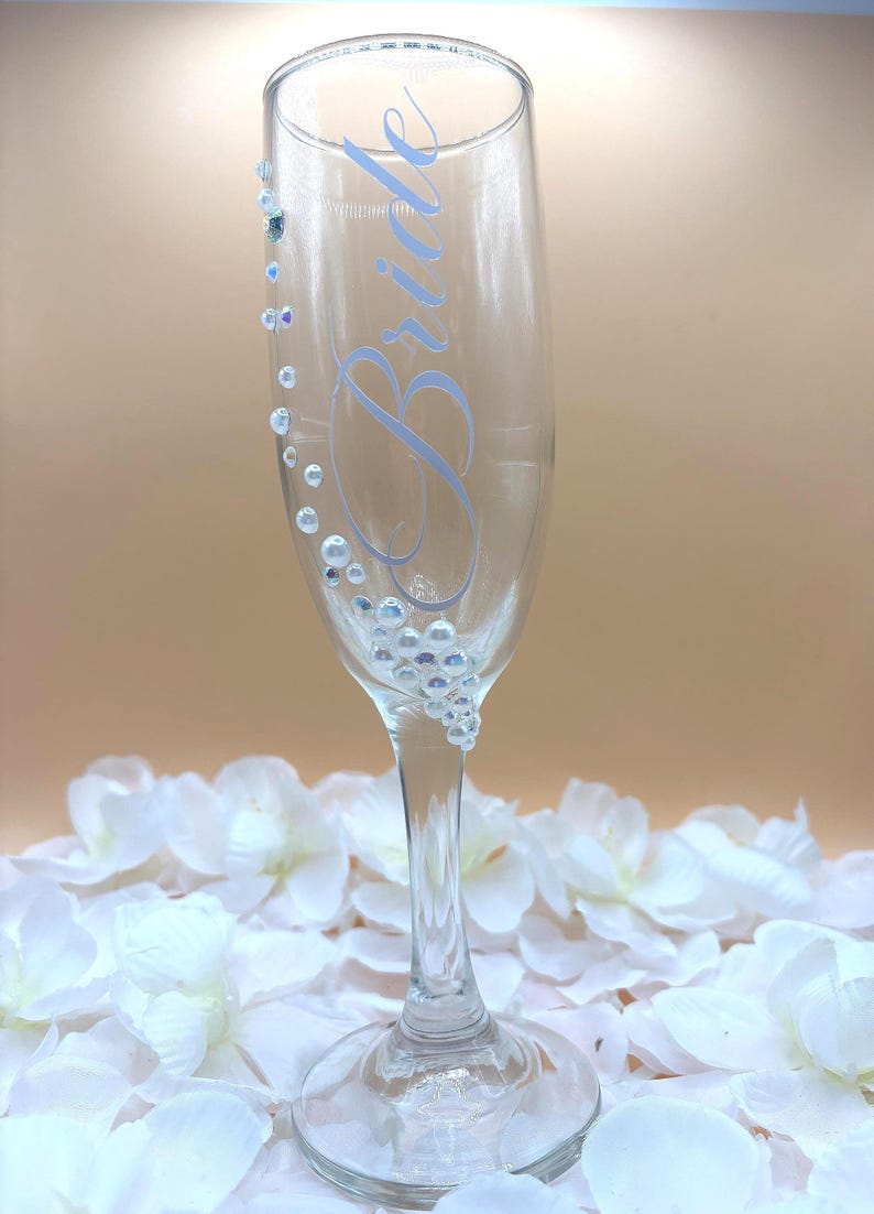 Puede incluir: Una copa de champ&aacute;n transparente con la palabra "Bride" en escritura azul claro. El vaso est&aacute; adornado con peque&ntilde;as cuentas claras e iridiscentes y cuentas m&aacute;s grandes con forma de perla. El vaso se apoya en un lecho de p&eacute;talos de flores blancas.