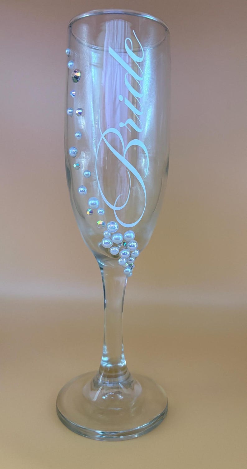 Puede incluir: Una copa de champ&aacute;n transparente con la palabra "Bride" en una elegante escritura. El cristal est&aacute; adornado con pedrer&iacute;a iridiscente y adornos perlados, a&ntilde;adiendo un toque de brillo. Perfecto para una boda o celebraci&oacute;n nupcial.