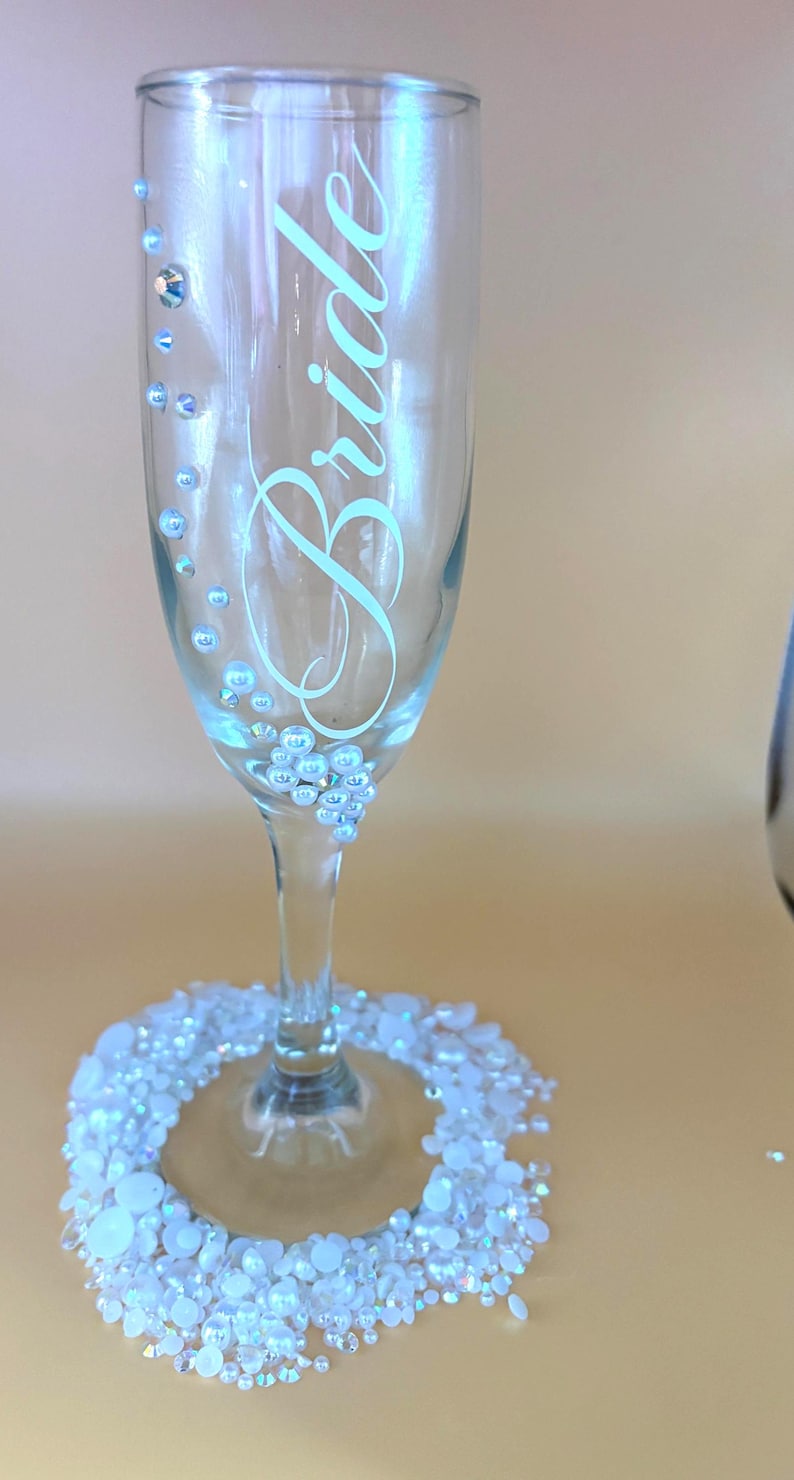 Puede incluir: Una copa de champ&aacute;n transparente con la palabra "Bride" en cursiva. El vaso est&aacute; adornado con pedrer&iacute;a iridiscente y una base de gemas blancas e iridiscentes. El dise&ntilde;o es elegante y adecuado para una boda o un evento nupcial.